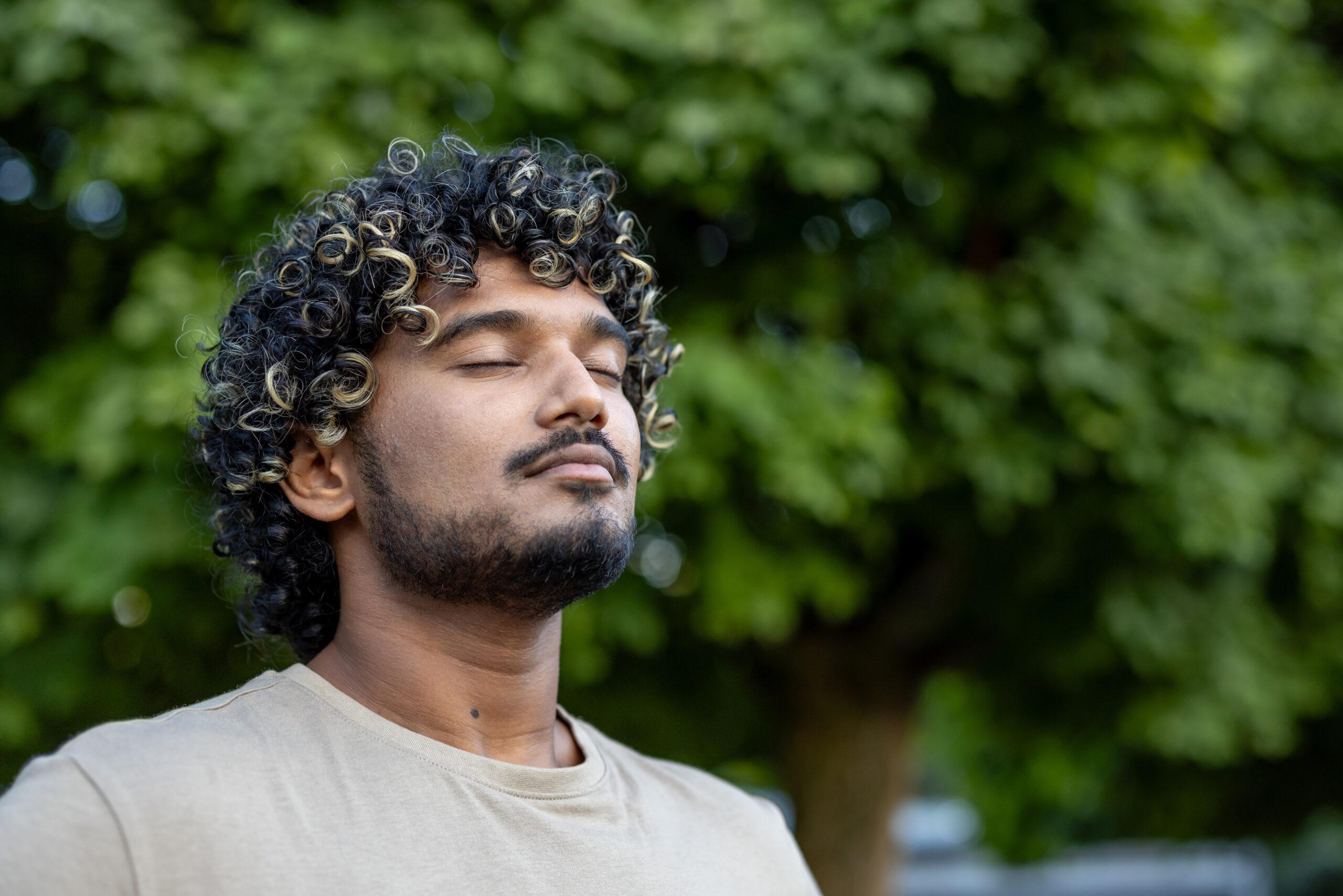A serene portrait of a man with curly hair enjoying a peaceful moment outside surrounded by green leaves, highlighting relaxation and mindfulness in nature.