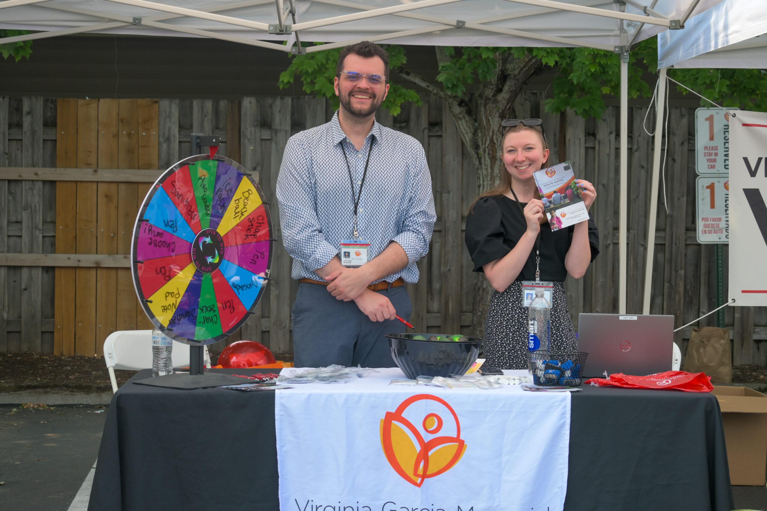 hombre y mujer de pie detras de un stand en una feria sosteniendo panfleto sobre virginia garcia 