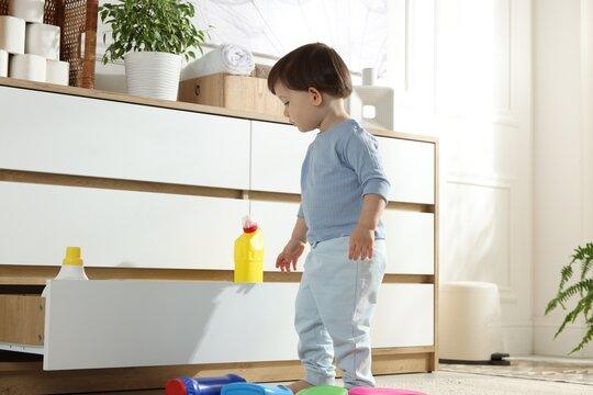 Little boy near cabinet with bottles of detergents at home