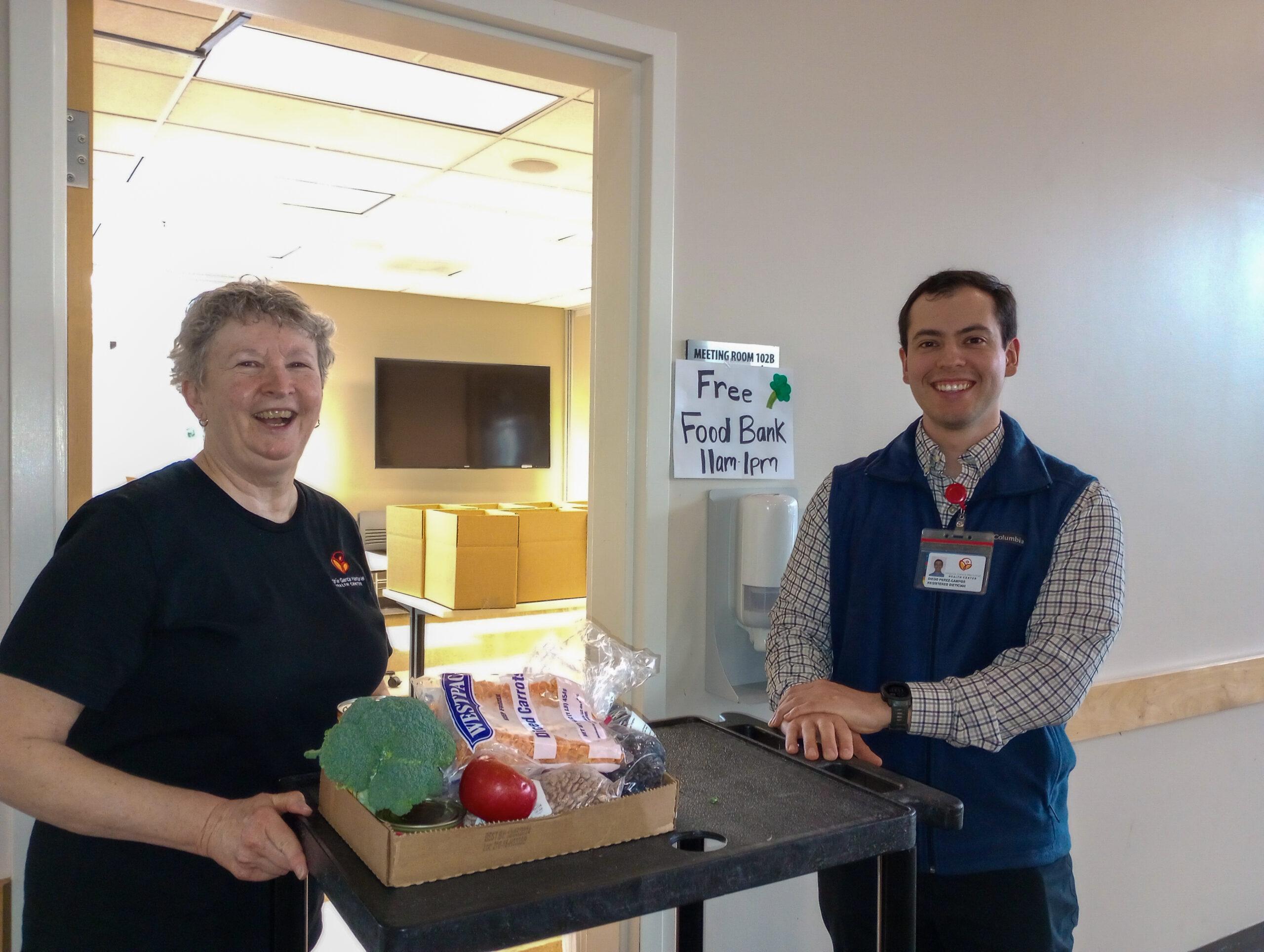 two volunteers are smiling in front of the foodbank with a basket of food