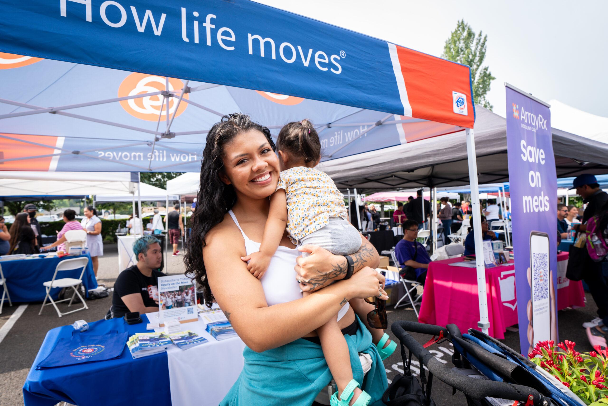 Una mujer con un niño en brazos sonríe delante de una caseta de una feria comunitaria