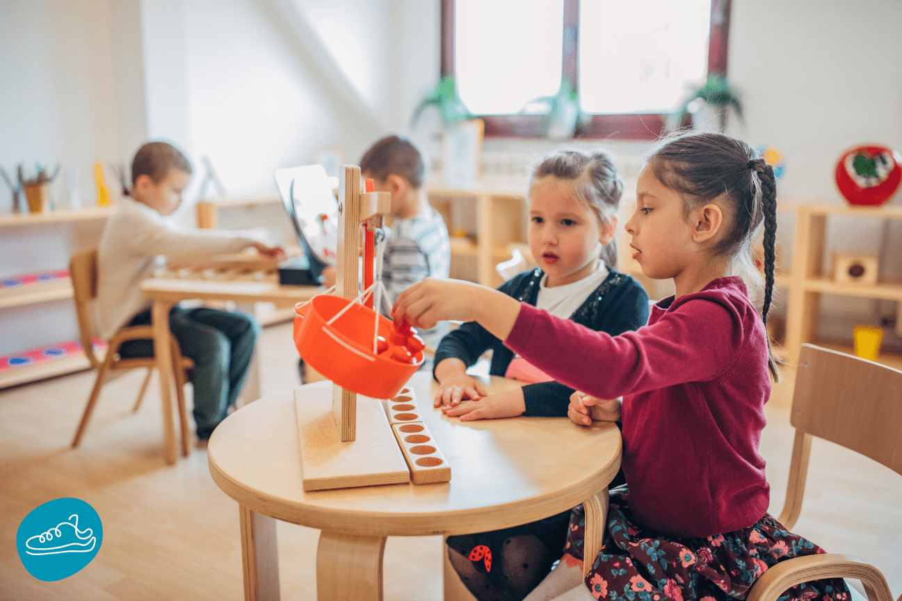 Two children are playing with a balance scale toy at a round table in a classroom. Another child is working at a desk in the background.