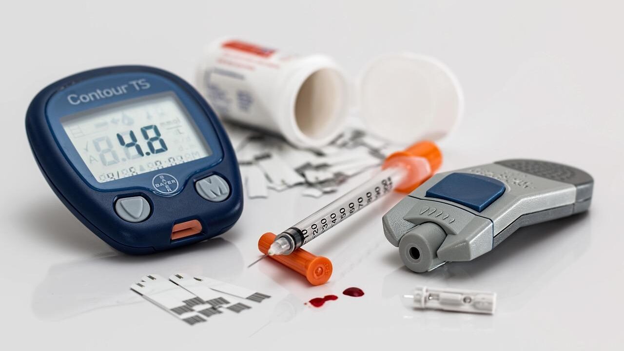 Un medidor de glucosa en sangre Bayer Contour TS que muestra una lectura de 14,8 junto a una jeringa de insulina, un dispositivo de punción, tiras reactivas y una gota de sangre sobre un fondo blanco